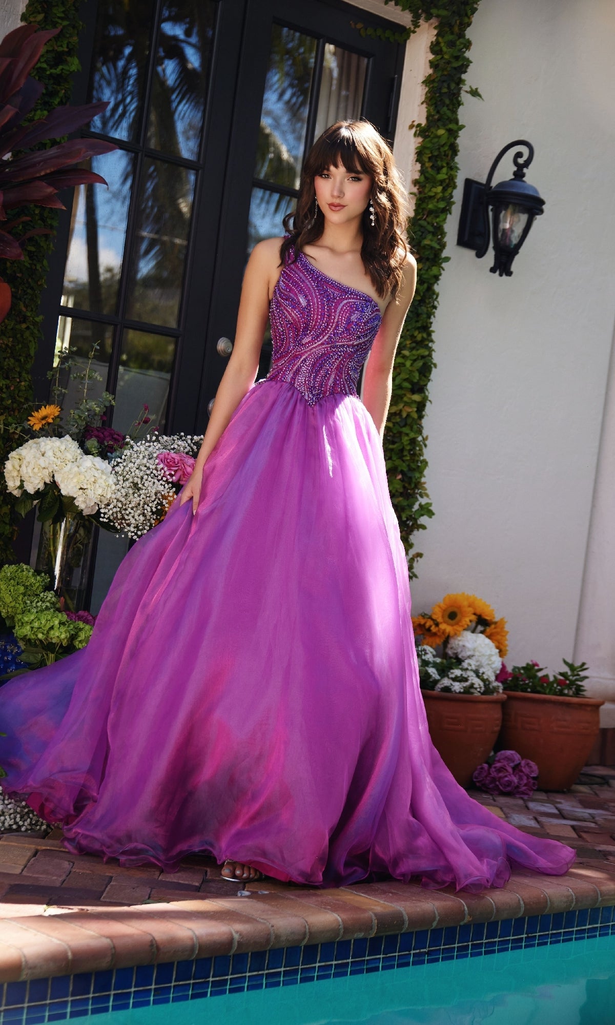 A woman in the Ava Presley 29258, a purple one-shoulder prom ball gown with a beaded bodice, stands by a pool amid potted plants and flowers near a glass door.